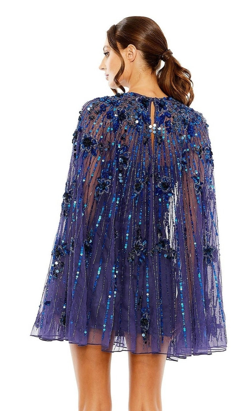 A woman with brown hair in a low ponytail is seen from the back wearing the Mac Duggal 5917 Beaded-Cape Short Hoco Dress, featuring a sheer blue sequin and floral-applique cape layered over a beaded cocktail dress.